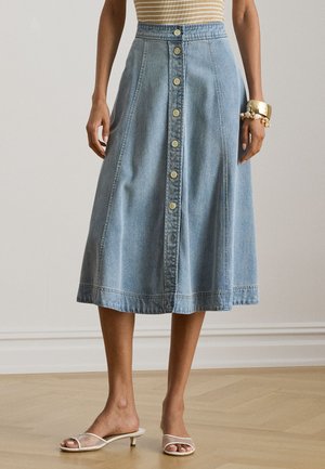 Femme portant une jupe en jean bleu mi-longue à boutons sur le devant, un haut beige rayé, des bracelets dorés, et des sandales blanches à talons bas sur un sol en bois.