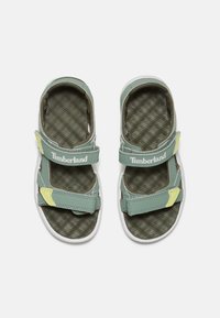 Groene synthetische sandalen met verstelbare klittenbandsluitingen, witte zolen en getextureerde bruine binnenzolen met een geruit patroon.