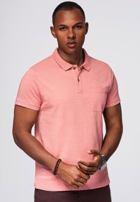 Svetle růžový polo tričko vyrobené z měkké látky, s límečkem, tříknoflíkovou légou a malou náprsní kapsou. Krátké rukávy a volný střih.