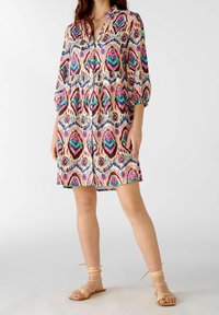 Vestido de patrón multicolor con cintura fruncida, mangas largas, cuello y corte por la rodilla; combinado con sandalias atadas de color beige claro.