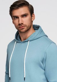 Jeune homme aux cheveux bruns courts et à la barbe, portant un sweat à capuche bleu clair avec des cordons blancs, regardant légèrement sur le côté.