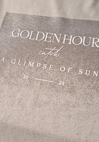 Kangas, jossa on harmaaseen gradienttiin vaihteleva tausta ja valkoiset tekstit, jotka lukevat "KULTAINEN HETKI, nappaa vilahdus auringosta 20 — 25." Pintakuvio on sileä.