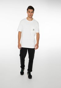 Witte katoenen t-shirt met een borstzak en een zwart logo. Gecombineerd met zwarte broek en donkere schoenen, staande tegen een lichte achtergrond.