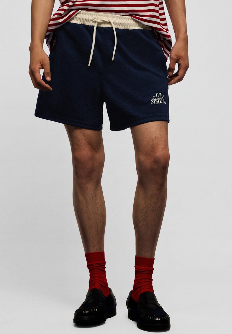 Homme portant un short bleu marine avec un cordon blanc, des chaussettes rouges, des chaussures noires et une chemise rayée rouge et blanche.