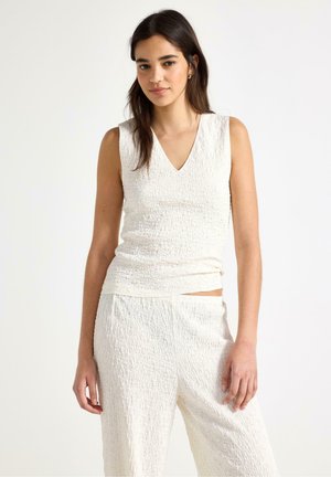 Pitkät mustat hiukset omaava nainen, joka pukeutuu kuvioituun valkoiseen hihattomaan toppiin ja vastaaviin leveälahkeisiin housuihin, seisoo yksinkertaisen taustan edessä.