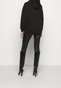 Svart oversized hoodie med huva, kombinerad med glänsande svarta läderleggings och knähöga stilettos, sedd från baksidan.