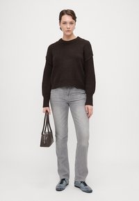 Smeđa pletena dukserica s dugim rukavima, uparena s svijetlosivim trapericama i plavim cipelama. Drži malu tamnu torbicu. Prikaz ležernog outfita.