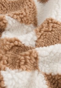 Braun-weiß karierter Stoff mit einer flauschigen, strukturierten Oberfläche. Das Muster zeigt abwechselnde Blöcke aus weichem, plüschigem Material.