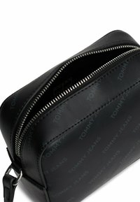Bolso cruzado de cuero negro con cierre de cremallera, diseño de logotipo en relieve "TOMMY JEANS" y detalles en metal plateado. Textura suave.