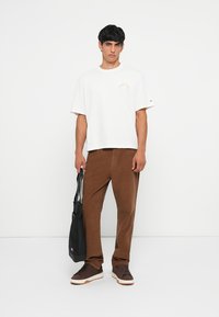Camiseta blanca de algodón con un pequeño gráfico, pantalones de pana marrones, bolsa negra y zapatillas marrones. Diseño simple con un corte relajado.