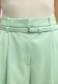 Myntgröna, vida byxor med en slät textur. Har ett bälte med en silverfärgad spänne och sidofickor, skräddarsydda för en avslappnad passform.