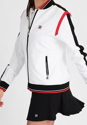 Weiße Jacke mit Reißverschluss mit roten und schwarzen Streifen an Ärmeln und Saum, kombiniert mit einem schwarzen Faltenrock mit Logo-Patch.