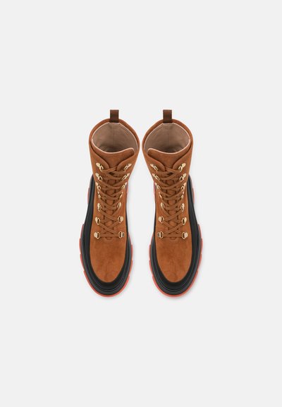 Brown suede ankle boots with gold metal eyelets and black rubber soles, featuring red accents and a pull tab at the back.