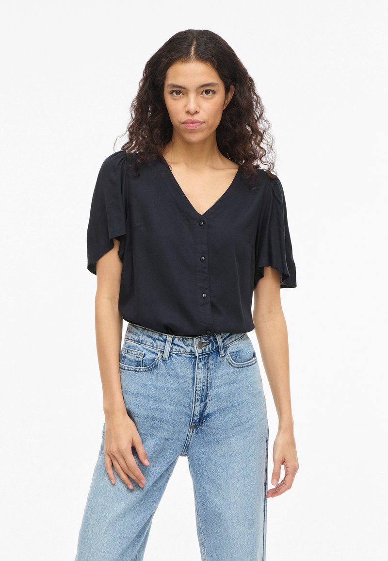 Kvinde med krøllet hår iført en sort V-halsbluse med knapper og lyseblå højtaljede jeans, stående mod en ensfarvet hvid baggrund.