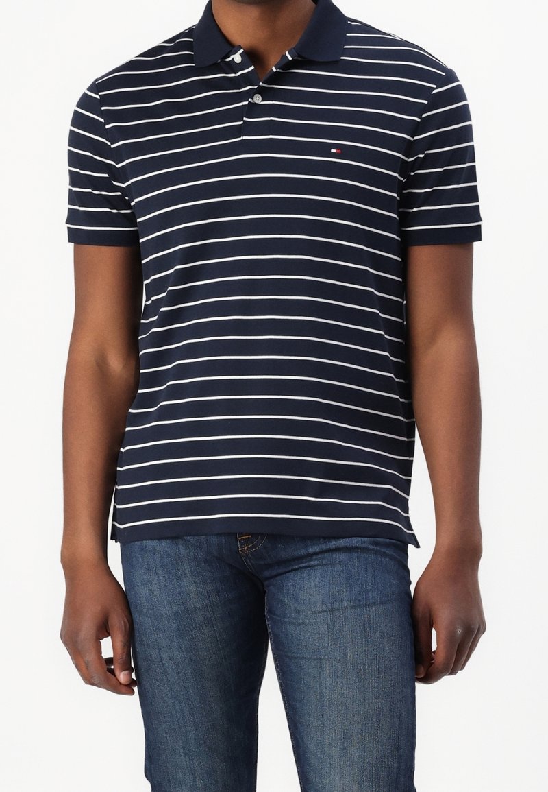 Homme portant un polo bleu marine à rayures horizontales blanches et un jean bleu, debout devant un fond blanc uni.