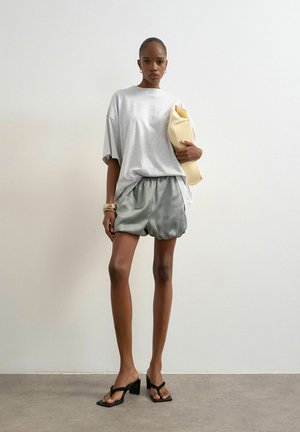 Woman wearing oversized light gray t-shirt, silky gray shorts, black heeled sandals, holding beige folded clutch bag against white wall.