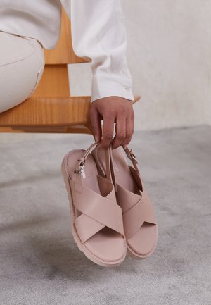 Main tenant une paire de sandales beiges à brides croisées avec boucles, posées sur une chaise en bois sur un sol gris clair.
