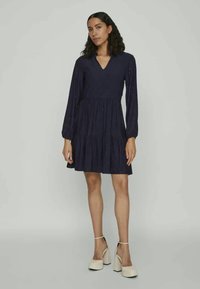 Ženska v temno modri obleki z dolgimi rokavi z izrezom in plastovitim krilom, obuta v bele visokolesne čevlje s trakovi okoli gležnjev.