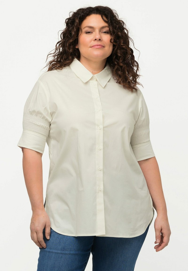 Helle beige Bluse mit Knöpfen, spitzem Kragen, kurzen Puffärmeln und texturierten Rüschen an den Bündchen. Glattes Material, lockere Passform.