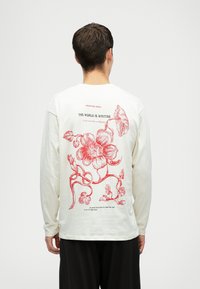 Person von hinten gesehen, der ein weißes Langarmshirt mit einem großen roten Blumenmuster und Text auf dem Rücken trägt.