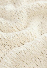 Szydełkowy koc w kremowym kolorze z teksturowanym wzorem. Miękka, gruba włóczka tworzy fale i głębię na powierzchni materiału.