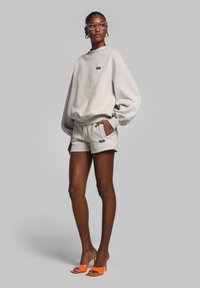 Lichtgrijze oversized sweatshirt en bijpassende shorts, met een klein zwart logo. Model draagt oranje hoge hakken en een paar transparante brillen.