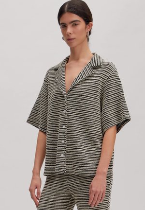 Frau trägt ein schwarz-weiß gestreiftes kurzärmeliges Hemd mit Knöpfen und passende Hose, steht vor einem schlichten hellen Hintergrund.