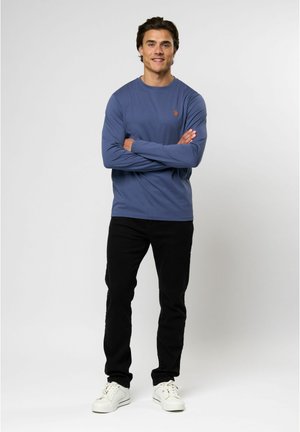 Zils krekls ar garām piedurknēm, izgatavots no mīksta auduma, ar mazu oranžu logotipu uz krūtīm, saskaņots ar melnām biksēm un baltām sporta kurpēm.