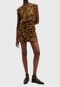 Kvinna klädd i en kort rynkad leopardmönstrad klänning med volangaxlar och svarta spetsiga platta skor med gyllene spännen.