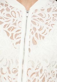 Veste blanche zippée en dentelle délicate avec un motif floral. Dotée d'un col blanc uni et d'une fermeture éclair visible, mettant en valeur une texture transparente.