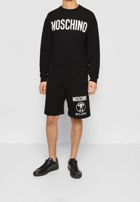 Černá mikina Moschino s bílým logem, spárovaná s černými kraťasy s logem a textem "Milano". V kombinaci s černými teniskami.