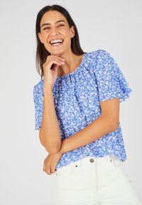 Blauwe bloemenblouse met korte fluttermouwen, gegatherde halslijn en witte bloemopdrukken. Gecombineerd met witte broek. Zacht materiaal.