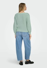 Femme vue de dos portant un pull tricoté vert menthe, un jean léger bleu ample et des chaussures en cuir noir sur un fond clair uni.