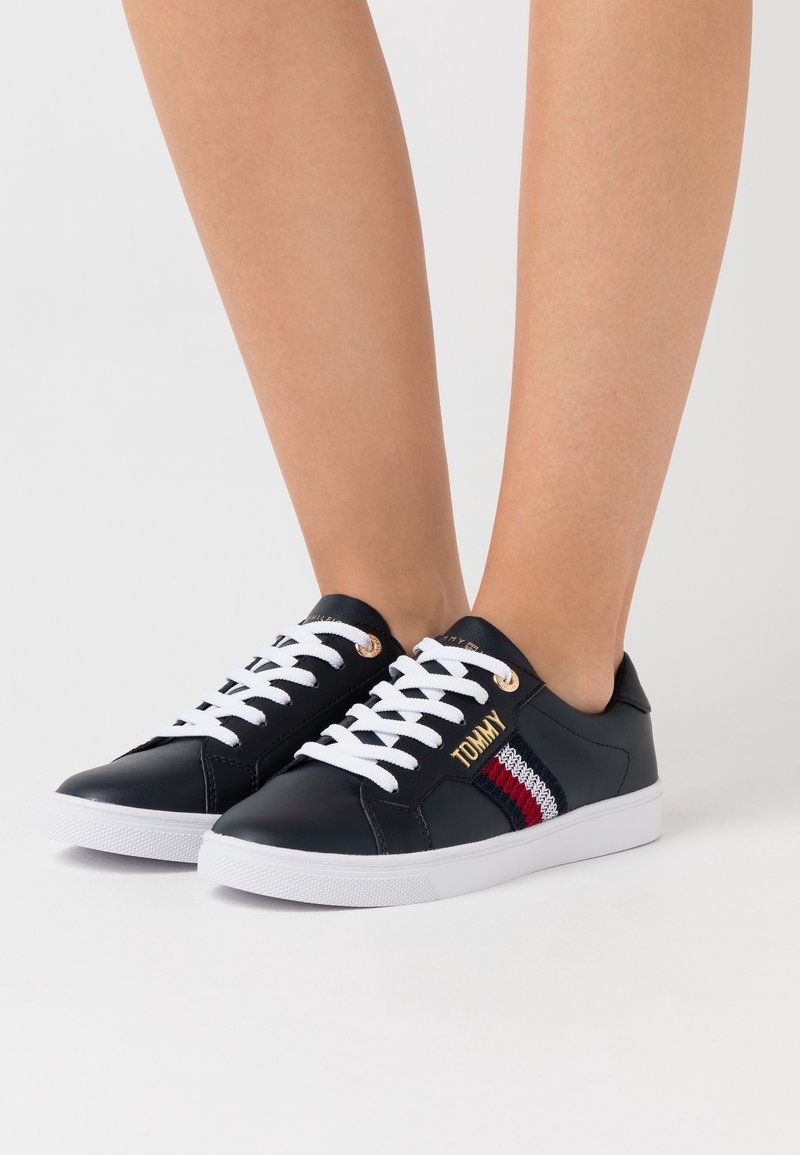 Baskets en cuir bleu marine avec lacets blancs, semelles blanches et bande en tissu rouge, blanc et bleu sur le côté, portées pieds nus.