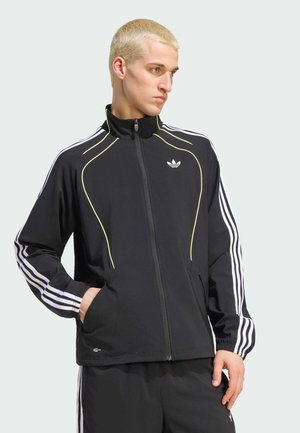 Mand med kort blondt hår iført en sort Adidas træningsjakke med hvide striber og gul kant, hænderne i lommerne, kigger til siden.