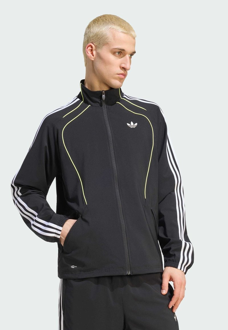Homme aux cheveux blonds courts portant une veste de survêtement noire Adidas avec des bandes blanches et un liseré jaune, mains dans les poches, regardant sur le côté.