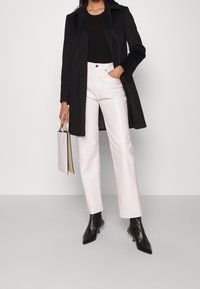 Abrigo negro sobre una blusa negra, pantalones blancos de cintura alta y botines negros. Sosteniendo un bolso pequeño de color claro.
