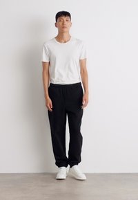 T-shirt blanc à manches courtes associé à un pantalon noir ample ; les chaussures sont des baskets blanches. L'arrière-plan est uni et de couleur claire.