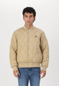 Chaqueta bomber acolchada beige con cierre de botones a presión, puños acanalados y un pequeño parche con el logo en el pecho izquierdo. Llevada sobre unos vaqueros azules.