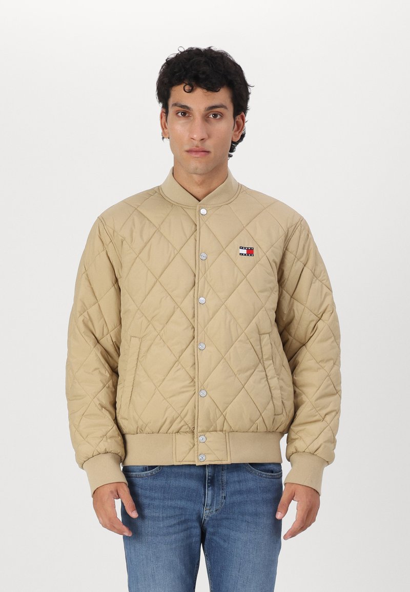 Chaqueta bomber acolchada beige con cierre de botones a presión, puños acanalados y un pequeño parche con el logo en el pecho izquierdo. Llevada sobre unos vaqueros azules.