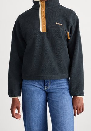 Sort fleece trøje med tan tryklåsning, lynlåslomme på brystet og blå jeans på en person mod en ensfarvet baggrund.