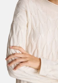 Krémový svetr s copánkovým vzorem, který se vyznačuje jemnou texturou, širokými žebrovanými manžetami a kulatým výstřihem. Ruka spočívající na rukávu.