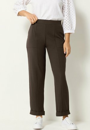 Mujer lleva pantalones rectos de pierna oscura con dobladillos remangados, combinados con una blusa blanca con ojales y zapatillas blancas, de pie frente a un fondo claro.