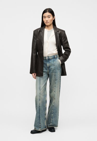 Γυναίκα με μακριά σκοτεινά μαλλιά, που φοράει μαύρο δερμάτινο σακάκι, εκρού ζακέτα, γκρι τοπ, φαρδιά ξεθωριασμένα τζιν παντελόνια και μαύρα παπούτσια με τετράγωνη μύτη, στεκόμενη με το χέρι στην τσέπη.