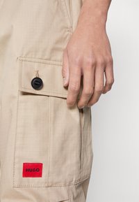 Beiga cargo-byxor med en texturerad tyg, sidficka med en svart knapp och en röd "HUGO"-etikett på fickan.