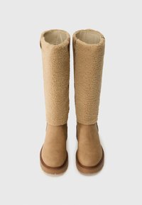 Botas marrones de altura hasta el tobillo con parte superior de shearling texturizado y sección inferior de cuero liso. Suela de goma plana, mostrando costuras a lo largo de los bordes.