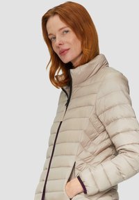 Beżowa kurtka puchowa z poziomymi i w kształcie zygzaka wzorami, wysoki kołnierz, zamek błyskawiczny z przodu oraz kontrastowe fioletowe akcenty na mankietach i zamku.