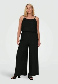 Mulher loira está de pé vestindo um top preto sem mangas e calças largas pretas, com saltos abertos pretos, contra um fundo simples.