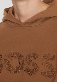 Gros plan sur l'encolure d'un sweat à capuche marron avec des lettres brodées texturées formant le mot "BOSS" sur la poitrine.