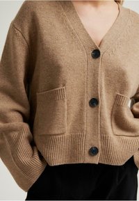 Beige kötött kardigán V-nyakkal, fekete gombokkal, bordás mandzsettákkal és két elülső zsebbel. Puha tapintás és laza szabás.